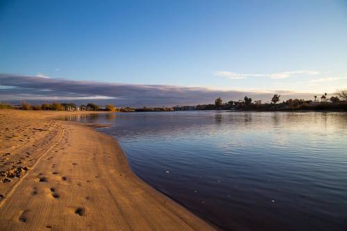 A serene riverbank at sunset, with gentle waves lapping against sandy shores and a clear sky above.