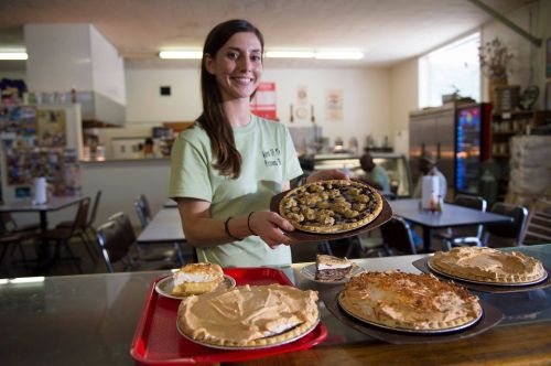 A smiling woman holds a pie while standing behind a counter with various baked goods on display.