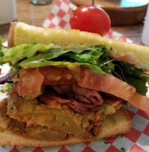 A close-up of a layered sandwich with lettuce, tomato, pickles, and sliced meats, topped with a cherry tomato.