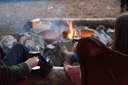 Two people sit by a campfire, holding glasses of red wine, enjoying a cozy outdoor evening.