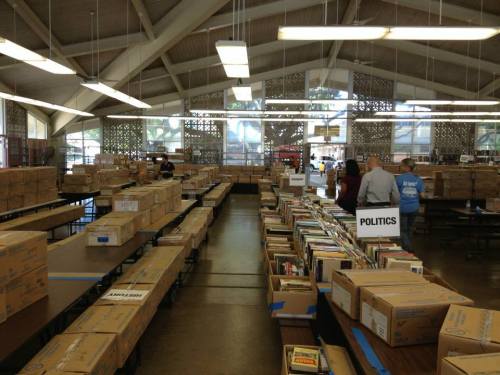 A spacious room filled with tables stacked with boxes and books, with people organizing items in a library setting.