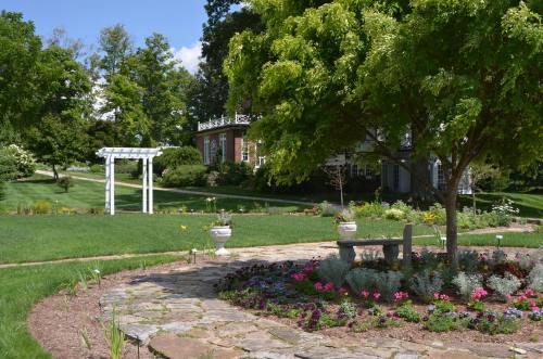 A serene garden with a stone path, colorful flowers, a bench, and a white arbor, surrounded by lush greenery and a house.