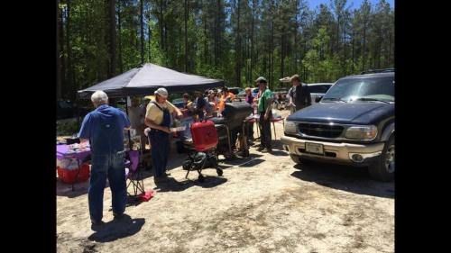 A group of people gather around a grill under a tent in a wooded area, enjoying a sunny day outdoors.