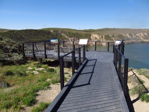A scenic overlook with a wooden deck, surrounded by green hills and a river, under a clear blue sky.