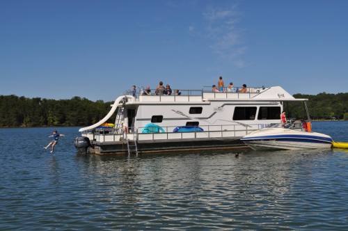 A houseboat on a lake with people on the deck and a speedboat nearby, under a clear blue sky.
