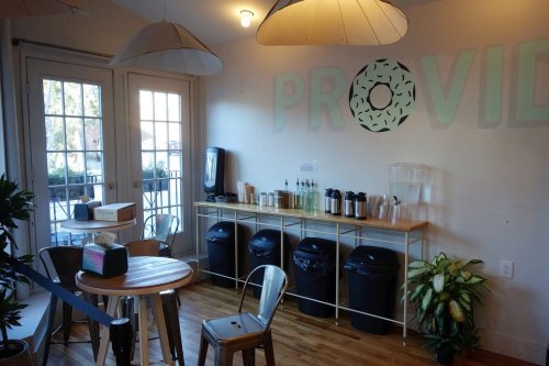 Cozy café interior with a table, chairs, and a counter featuring drinks and trash bins, plus large windows.