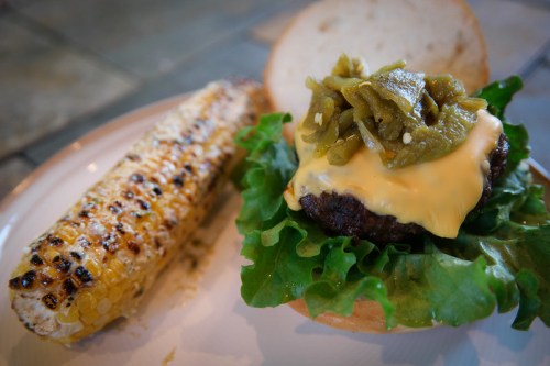 A juicy burger with melted cheese, lettuce, and green toppings, served with a grilled corn on the side.