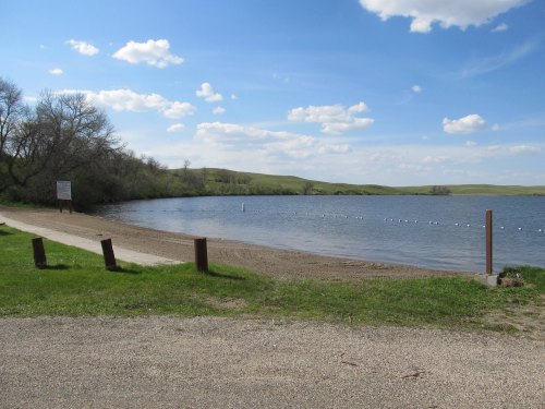 A serene lakeside view with a sandy beach, grassy area, and blue sky dotted with clouds.