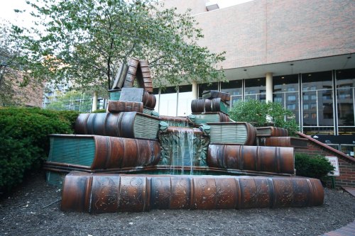 A whimsical sculpture of stacked books with a small waterfall, surrounded by greenery and a brick pathway.