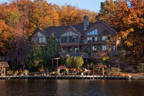 A large wooden house with multiple balconies, surrounded by autumn trees, overlooking a calm lake and a dock.