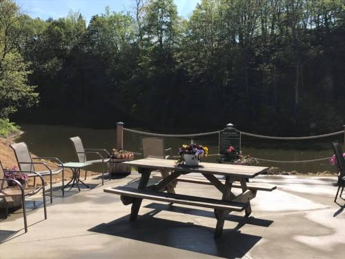 A serene lakeside view with a picnic table, chairs, and colorful flowers, surrounded by trees and calm water.