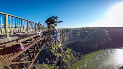 A person jumps off a bridge with a parachute, overlooking a river and rocky cliffs under a clear blue sky.