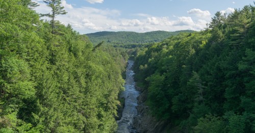 A scenic view of a river winding through lush green forests and rolling hills under a blue sky with fluffy clouds.