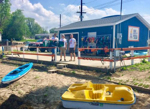 Three people stand in front of a rental shop with kayaks and paddleboards, surrounded by sandy ground and parked vehicles.