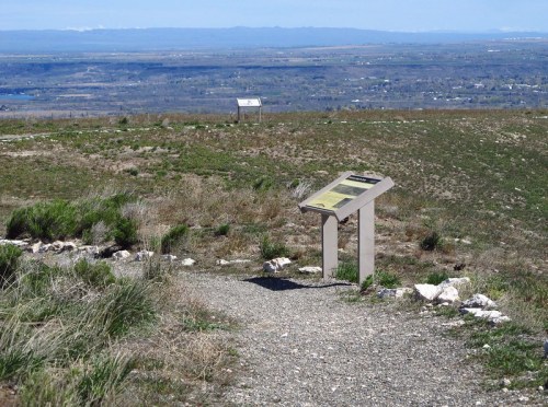 A viewpoint sign on a grassy hilltop, overlooking a valley and distant mountains under a clear blue sky.
