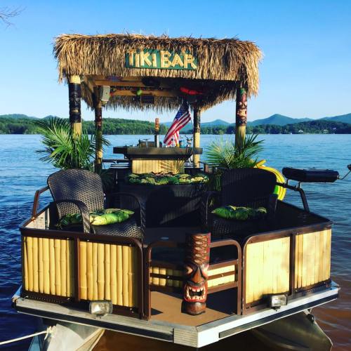 A tiki bar on a boat, featuring bamboo decor, tropical plants, and a scenic lake view with mountains in the background.