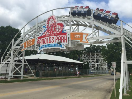 Welcome sign at Arnolds Park Amusement Park, featuring a roller coaster and a cloudy sky in the background.