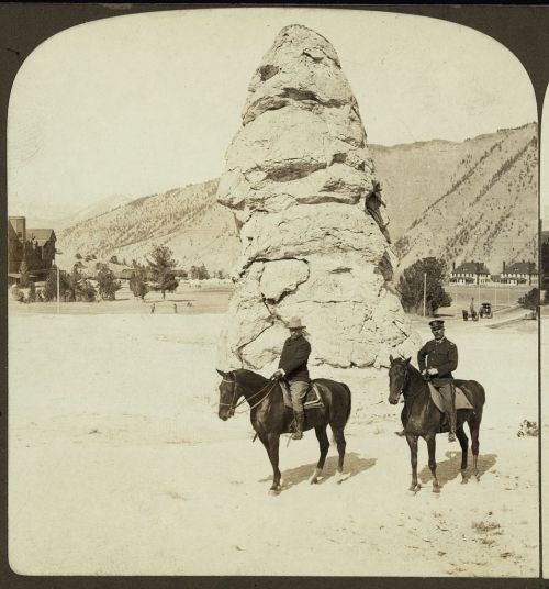 Two men on horseback pose in front of a large rock formation, with mountains and buildings in the background.