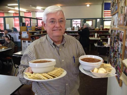 A smiling man holds two bowls of chili and plates of crackers in a dining area with a patriotic backdrop.