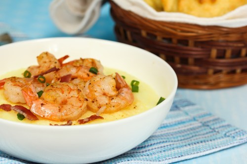 A bowl of shrimp and grits topped with green onions and bacon, with a basket of bread in the background.