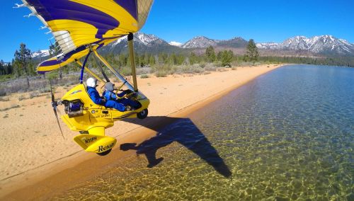 A yellow ultralight aircraft flying over a sandy beach and clear water, with mountains in the background under a blue sky.