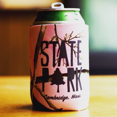 A green can in a pink koozie with "STATE PARK" and "Cambridge, Mass" printed on it, resting on a wooden surface.