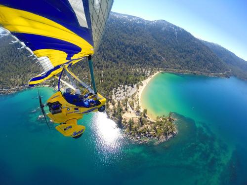 A colorful ultralight aircraft flying over a scenic lake with a sandy beach and lush green forests in the background.