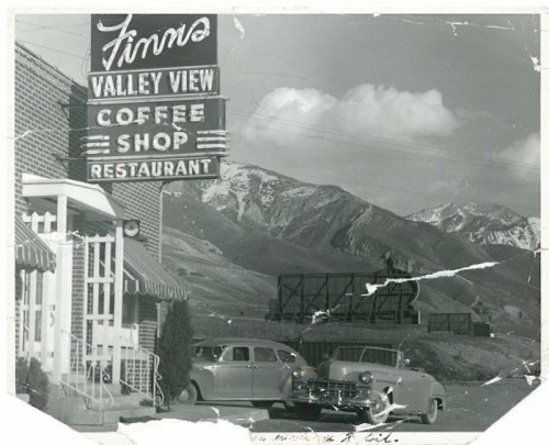 Black and white photo of a coffee shop sign with mountains in the background and vintage cars parked outside.