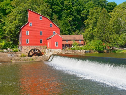 A red historic mill beside a calm river, with a waterfall cascading over a stone dam and lush greenery in the background.