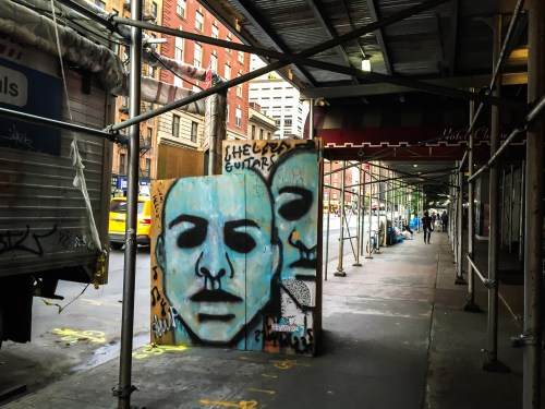 Street scene featuring graffiti of two faces on a barrier, with scaffolding and urban buildings in the background.