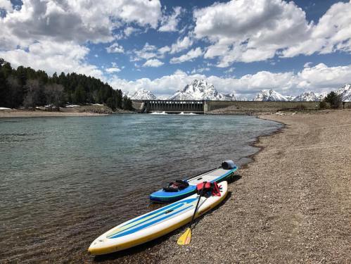 Two paddleboards on a sandy shore by a river, with mountains and a dam in the background under a partly cloudy sky.