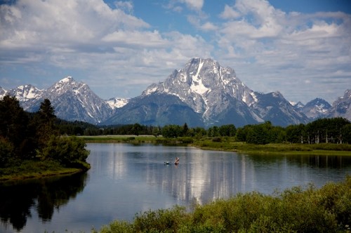 A serene lake reflects snow-capped mountains under a blue sky, with two people kayaking in the foreground.