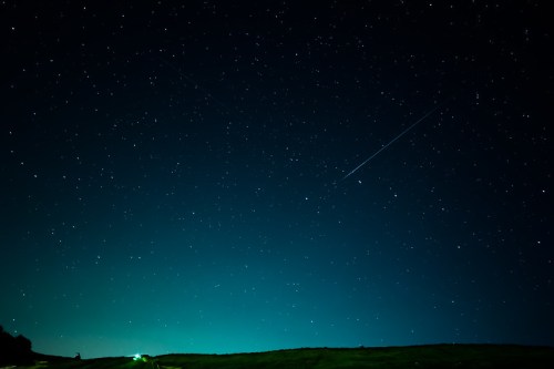 A clear night sky filled with stars, featuring a bright meteor streaking across the horizon.