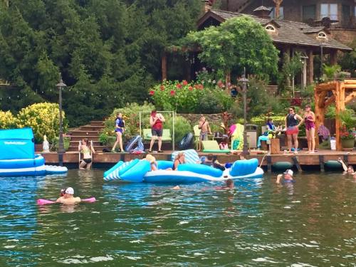 A lively lakeside scene with people swimming, lounging on inflatables, and enjoying a sunny day by a garden and wooden deck.