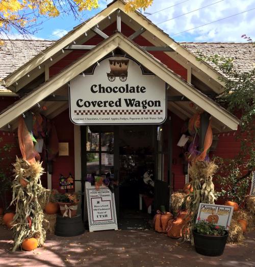 A red building with a sign reading "Chocolate Covered Wagon," decorated for fall with pumpkins and scarecrows.