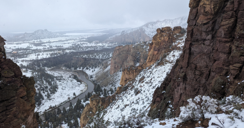 Snow-covered cliffs overlook a winding river and valley, with a cloudy sky in the background.