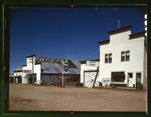 A dusty street scene featuring buildings with signs for "Wisdom" and "Baker's Garage" under a clear blue sky.