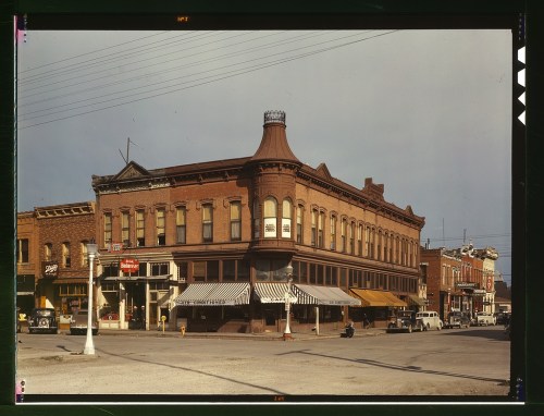 Historic brick building with a decorative turret, storefronts, and vintage cars in a small town setting.