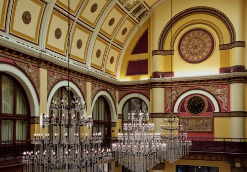 Elegant interior of a grand hall featuring ornate chandeliers, decorative arches, and a large clock on the wall.