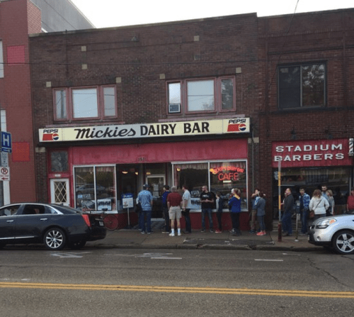 A busy street scene featuring Mickie's Dairy Bar with a line of customers outside and nearby barbershop.