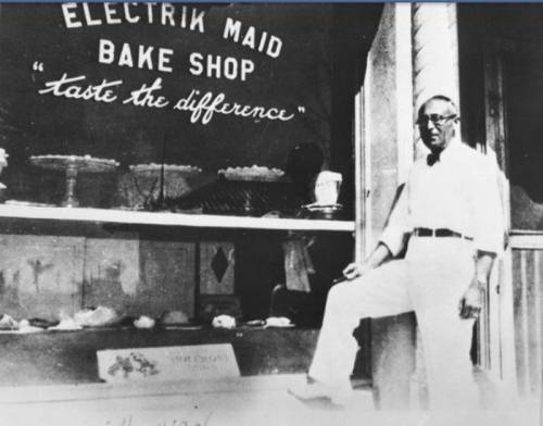 A man stands outside the Electrik Maid Bake Shop, showcasing baked goods in the window with a sign that reads "taste the difference."