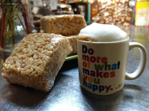 A colorful mug with foam coffee beside wrapped rice crispy treats on a table.