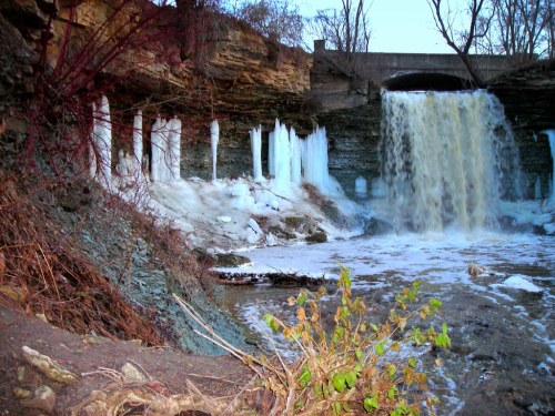 A waterfall cascades over rocky cliffs, surrounded by icicles and winter vegetation.