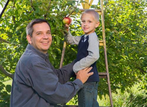A man helps a young boy pick an apple from a tree while standing on a ladder in a sunny orchard.