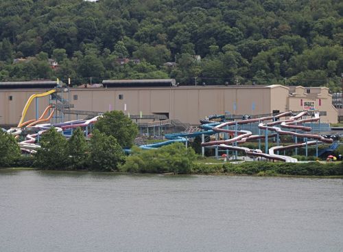 A water park with colorful slides beside a river, surrounded by green trees and a large building in the background.