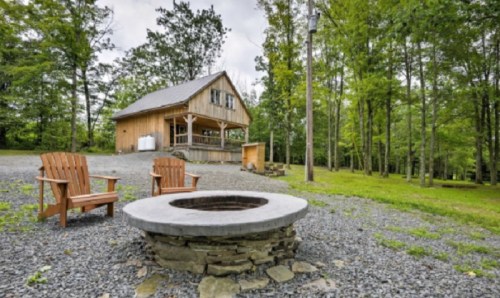A rustic cabin surrounded by trees, featuring a fire pit and two wooden chairs in a serene outdoor setting.