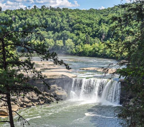 A scenic waterfall cascading into a river, surrounded by lush green trees and rocky formations under a blue sky.