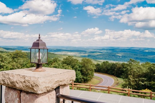 A lantern on a stone pillar overlooks a scenic valley and winding road under a blue sky with fluffy clouds.
