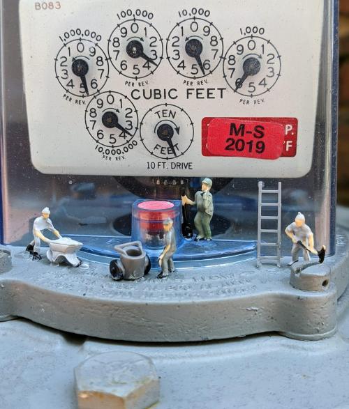 A close-up of a gas meter with miniature construction figures working around it, including a wheelbarrow and a ladder.