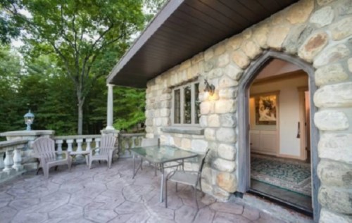 A stone patio with a table and chairs, leading to an arched doorway and surrounded by lush greenery.